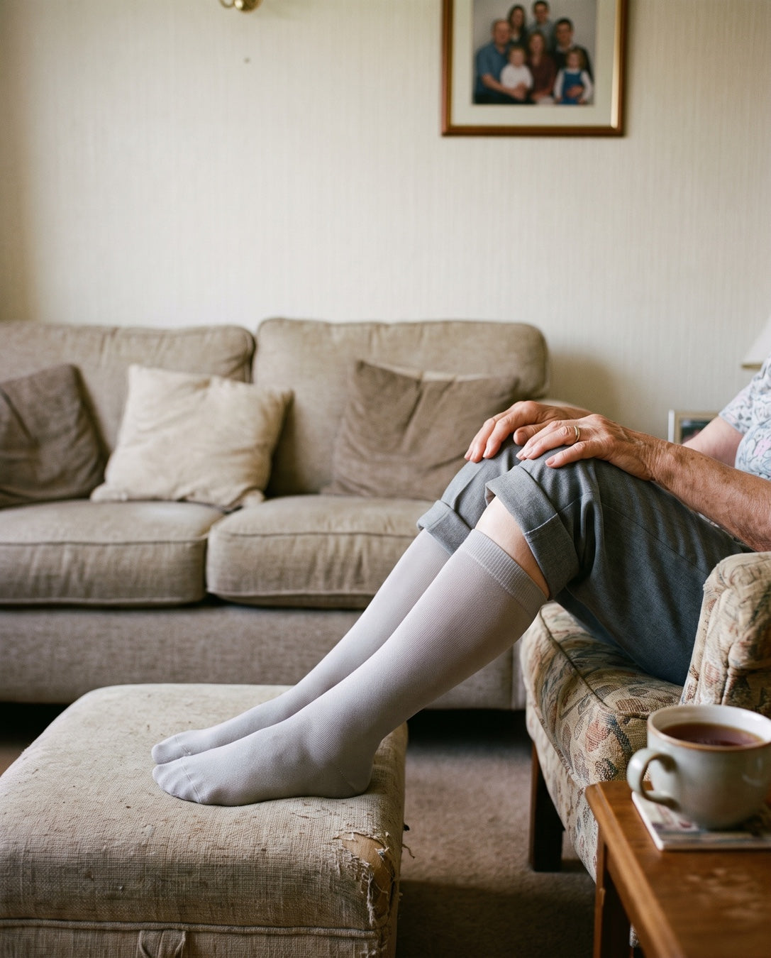 A woman in her seventies resting on her sofa with a cup of tea, wearing grey knee-high compression socks