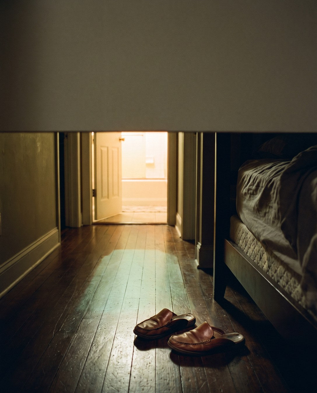 A dark hallway at night with the bathroom light on at the far end and slippers kicked off by the bed