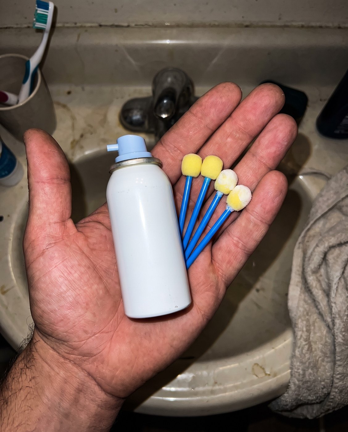 A hand holding a home freeze spray and four foam applicators over a bathroom sink