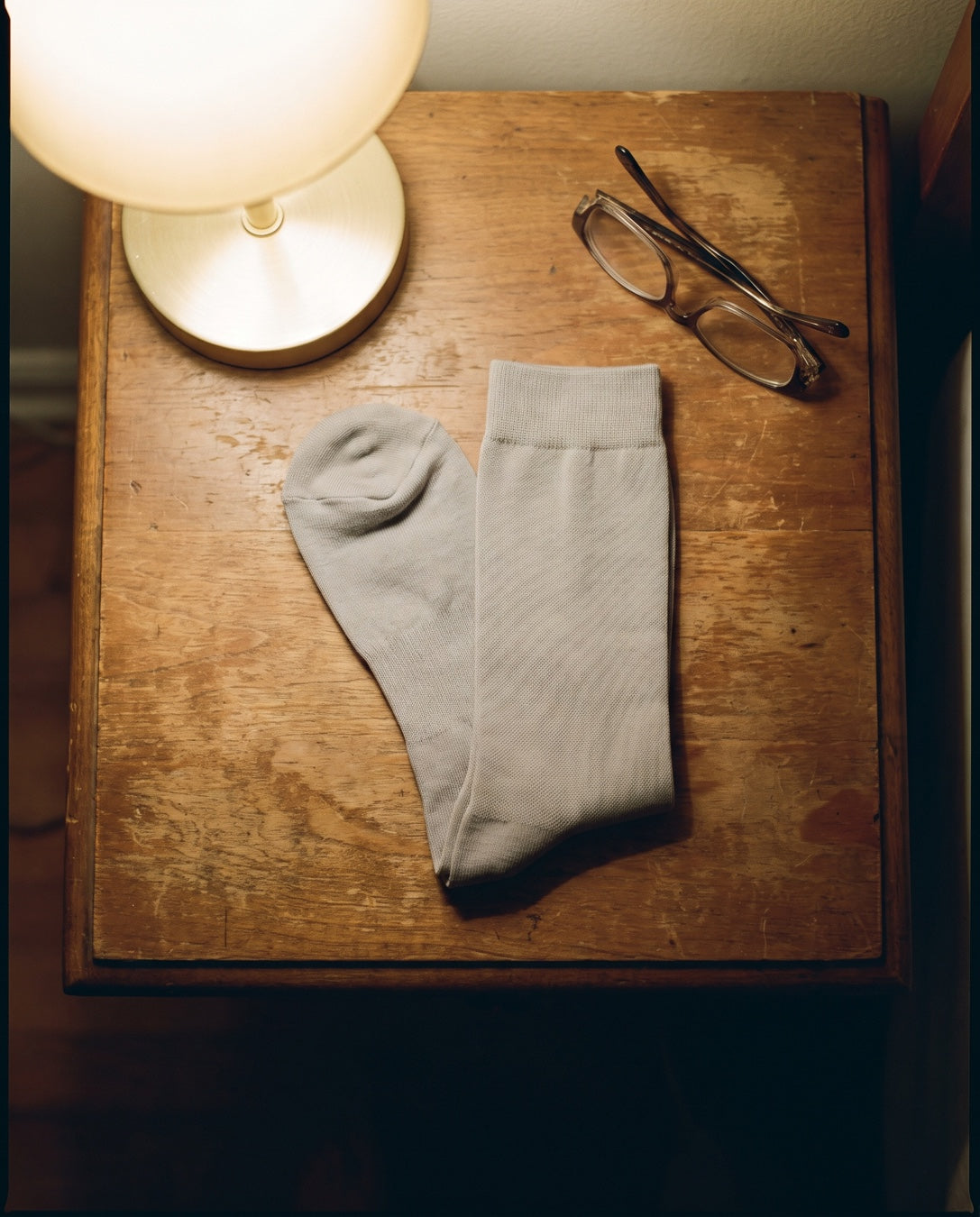 A pair of grey compression socks folded on a nightstand next to reading glasses and a lamp