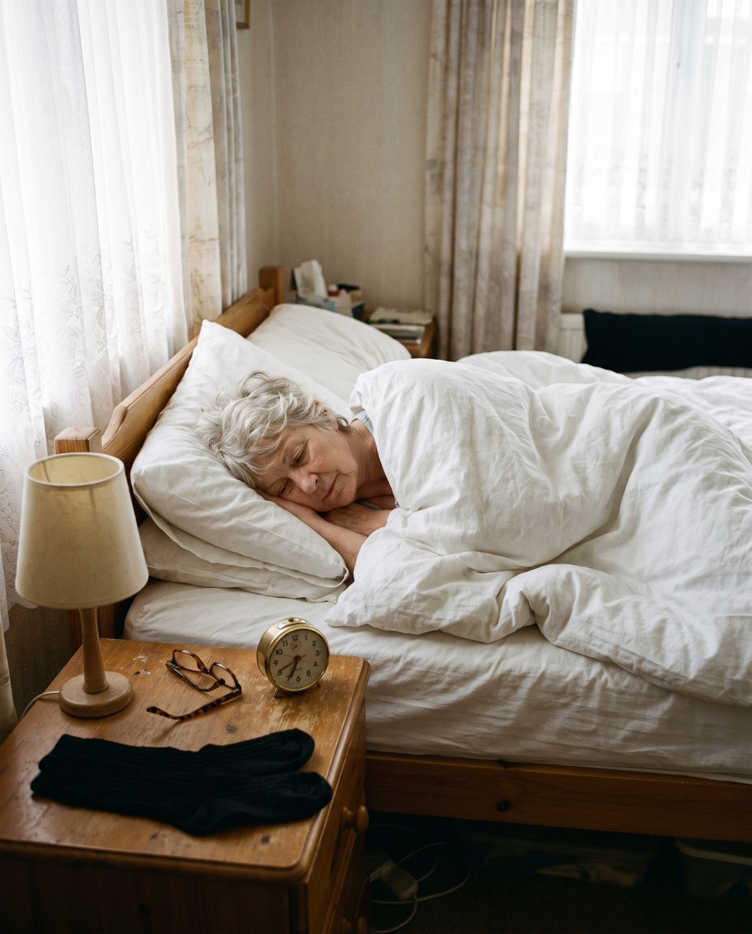 A woman in her sixties asleep in bed in the morning light, her socks folded on the nightstand