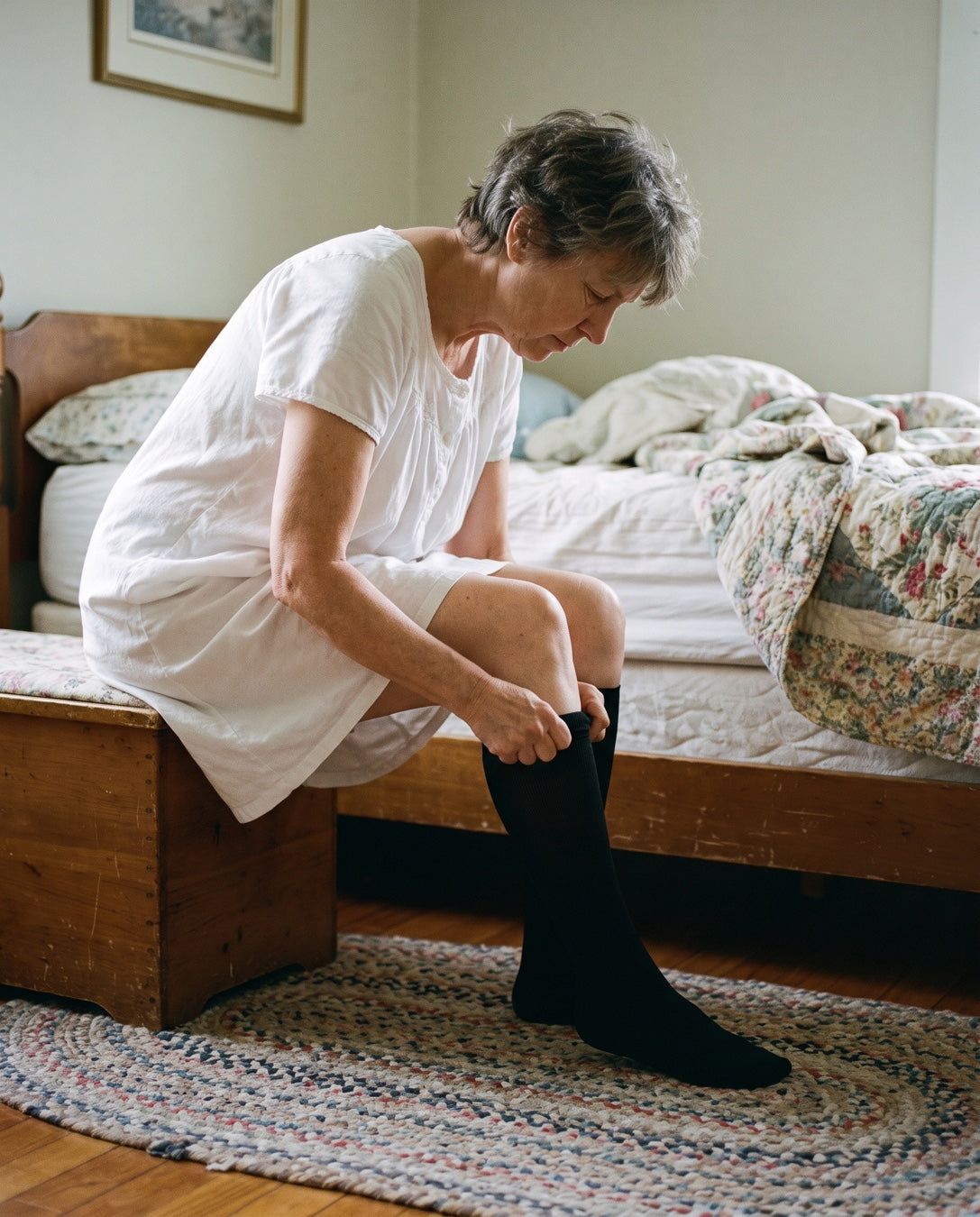 A woman in her sixties sitting on the edge of her bed in the morning, pulling a knee-high compression sock up her calf